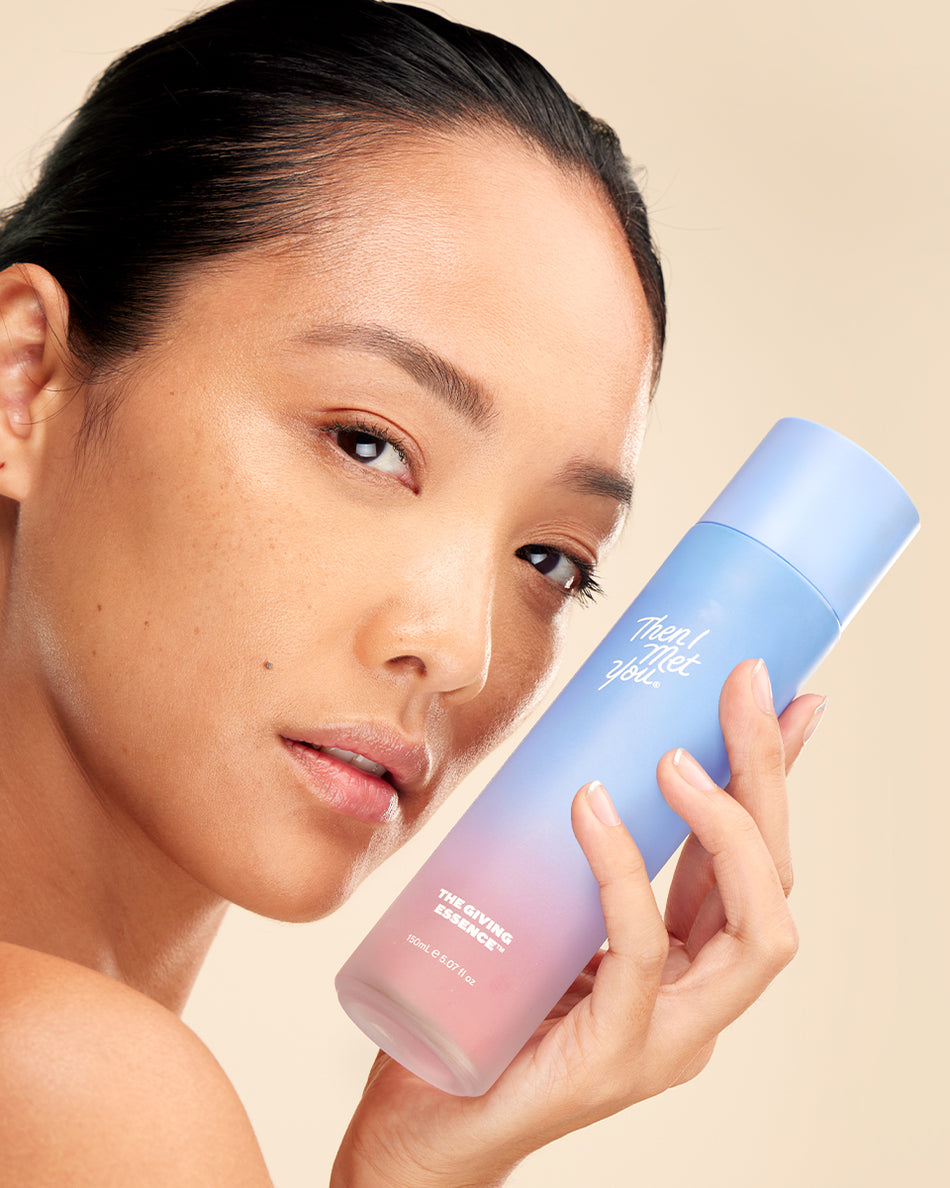 Woman holding a gradient blue to pink frosted bottle of The Giving Essence skincare product close to her face.